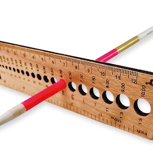 May include: A wooden knitting needle gauge with a ruler marked in inches and 16ths of an inch. The gauge has a series of holes for measuring knitting needles. A knitting needle is placed through one of the holes.