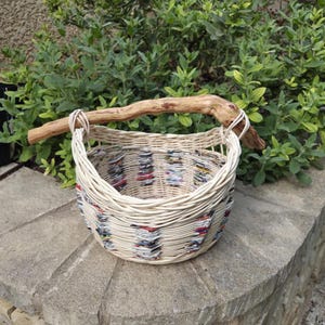 Peut inclure: Un panier rond tressé en osier clair et en bandes de papier recyclé coloré. Le panier est doté d'une poignée robuste en branche de bois naturel, fixée solidement au bord tressé de chaque côté de l'ouverture.