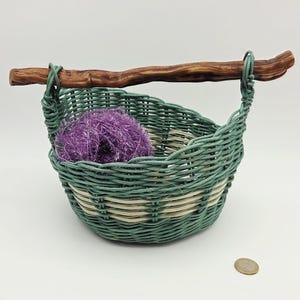 Peut inclure: Un panier tissé à la main avec un motif vert et beige, doté d'une poignée en bois. À l'intérieur du panier se trouve une boule de fil violet. Le panier est rond et il y a une pièce de monnaie à côté.