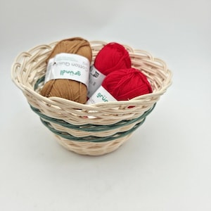 Peut inclure: Un panier en osier tressé avec des brins beige clair et vert foncé contenant une pelote de laine beige et deux pelotes de laine rouge, chacune avec une étiquette blanche indiquant Gründl Cotton Quick uni, disposées dans le récipient rond.