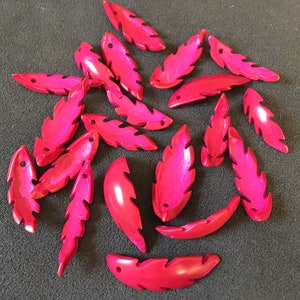 Puede incluir: Una colección de 18 cuentas de color rosa brillante en forma de hoja con un acabado brillante. Las cuentas están hechas de un material liso y duro y tienen un agujero perforado en la parte superior para enhebrar.