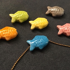 Peut inclure: Six poissons en céramique de différentes couleurs, dont jaune, orange, rose, bleu, vert et gris. Les poissons ont tous une forme similaire, avec un corps rond et une queue pointue. Ils sont disposés sur une surface gris foncé.