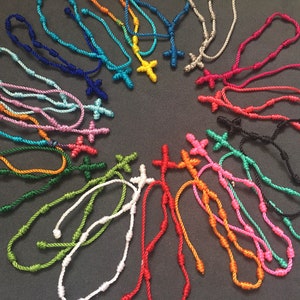 Puede incluir: Una colección de pulseras de cuerda anudada de colores con un dije de cruz. Cada pulsera tiene un color diferente y presenta un pequeño dije de cruz.