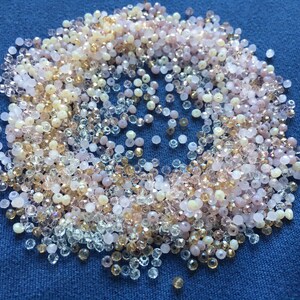May include: A large assortment of small, clear glass beads in various shades of pink, beige, and white. The beads are scattered on a blue surface.