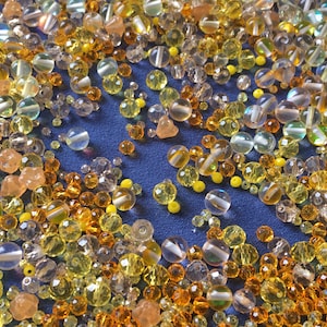 May include: A collection of assorted glass beads in various shades of yellow, orange, and clear. The beads are scattered on a blue surface.