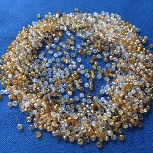 May include: A close-up of a large pile of clear and gold glass beads. The beads are small and round, and they are scattered on a blue surface.