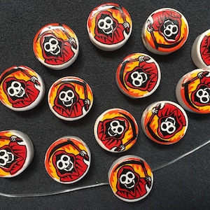 May include: Twelve round white ceramic buttons featuring a black and red image of a skeleton wearing a red hooded robe and holding a scythe. The skeleton has three eyes and is surrounded by flames.