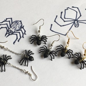 Peut inclure: Six paires de boucles d'oreilles en forme d'araignée noires avec des crochets argentés et dorés. Certaines boucles d'oreilles ont une chaîne reliant l'araignée au crochet.