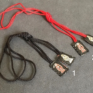 Peut inclure: Deux colliers avec de petits pendentifs rectangulaires. L'un a un cordon rouge, l'autre un cordon noir. Chaque pendentif est noir avec un motif brodé blanc et rouge. Les pendentifs sont numérotés 1 et 2.