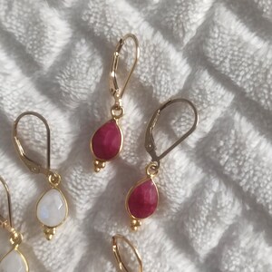 Peut inclure: Une collection de boucles d'oreilles pendantes avec des crochets dormeuses dorés et des pierres en forme de goutte. L'ensemble comprend des paires avec des pierres blanches translucides et des pierres rose foncé, ornées de trois petites perles dorées.