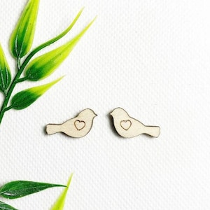 Op de afbeelding: Twee kleine houten vogeluitsparingen met hartvormige details. De vogels zijn lichtbruin en staan tegen een witte achtergrond. Groene bladeren bevinden zich in de linkerbovenhoek. De vogels zijn een knutsel- of doe-het-zelf-artikel.