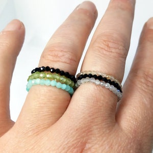 Puede incluir: Primer plano de varios anillos de cuentas en varios colores, incluyendo negro, verde y azul claro. Los anillos se llevan en los dedos, mostrando su delicado diseño. Las cuentas parecen pequeñas y facetadas, creando un efecto brillante.