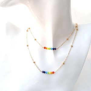 Peut inclure: Collier à deux rangs avec une chaîne dorée et des perles colorées arc-en-ciel. Le collier est présenté sur un mannequin blanc. Les perles sont disposées en dégradé de couleurs, créant un design vibrant et accrocheur.
