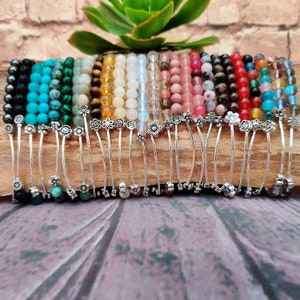 Può includere: Una collezione di 24 braccialetti colorati con perline e ciondoli a forma di fiore argentati. Ogni braccialetto è realizzato con diversi tipi di perline, tra cui turchese, verde, giallo, bianco, rosa, rosso e blu.