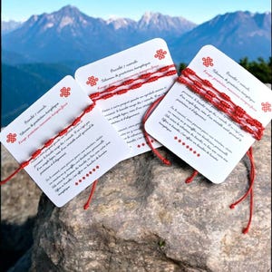 Op de afbeelding: Drie witte kaarten met rode touw armbanden, elk met tekst en een rood knoopsymbool. De kaarten liggen op een grijze rots met een bergketen op de achtergrond. De armbanden zijn een beschermingstalisman.