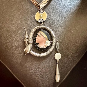 May include: A vintage necklace with a silver chain and pendant. The pendant features a woman's profile, a snake, and a dangling earring. The necklace has a cream-coloured background and black accents.