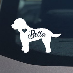 May include: White vinyl decal of a dog with the name "Bella" in script font. A black heart is next to the dog's head. The decal is adhered to a car window.