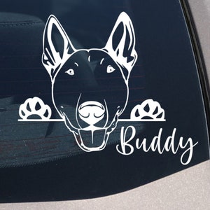 Puede incluir: Calcomanía de vinilo blanco para coche con la cara de un perro y el nombre "Buddy" en cursiva. La cabeza del perro está sobre una línea, con huellas de patas a cada lado. Adecuado para aplicar en la ventana de un coche, un accesorio temático de mascotas.