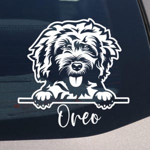 Puede incluir: Adhesivo de vinilo blanco con la cara de un perro y el nombre "Oreo" debajo. El perro tiene pelaje esponjoso y una expresión feliz, con la lengua fuera. Diseñado para ser aplicado en la ventana de un coche.