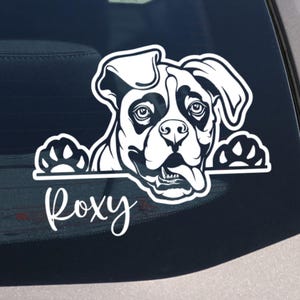 Op de afbeelding: Een zwart-witte hondensticker met de naam "Roxy" in cursief schrift. Het gezicht en de poten van de hond zijn zichtbaar, met zijn tong uitgestoken. De sticker zit op een autoruit.