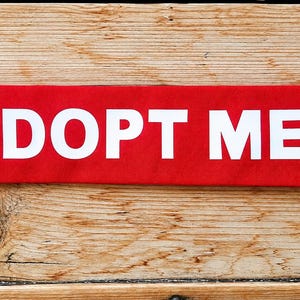 May include: A rectangular red fabric band featuring the white text ADOPT ME in capital letters followed by a white paw print graphic. The band is designed for display and is presented as a flat, horizontal strip with clean, straight edges.