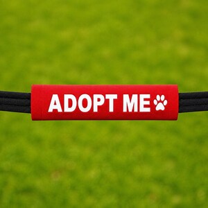 May include: A rectangular red leash sleeve featuring the white text ADOPT ME and a white paw print icon. The sleeve is attached to a black leash, providing a clear message for pet visibility during walks or public outings with a rescue animal.