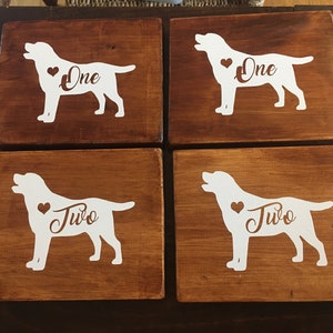 Op de afbeelding: Vier houten onderzetters met een donkerbruine afwerking. Elke onderzetter heeft een wit silhouet van een hond met een hart en het nummer "Een" of "Twee" in een decoratief lettertype. Een charmante toevoeging aan elk huis.