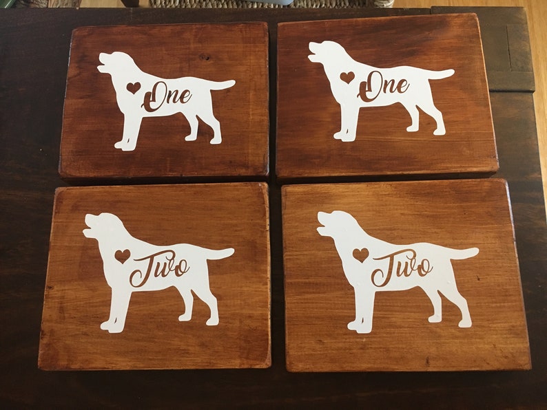 Op de afbeelding: Vier houten onderzetters met een donkerbruine afwerking. Elke onderzetter heeft een wit silhouet van een hond met een hart en het nummer "Een" of "Twee" in een decoratief lettertype. Een charmante toevoeging aan elk huis.