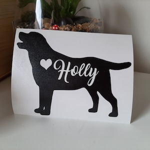 Op de afbeelding: Zwart Labrador Retriever silhouet met een wit hart en de naam "Holly" in een sierlijk lettertype. De sticker staat op een witte achtergrond. De hond kijkt naar links met zijn mond open.