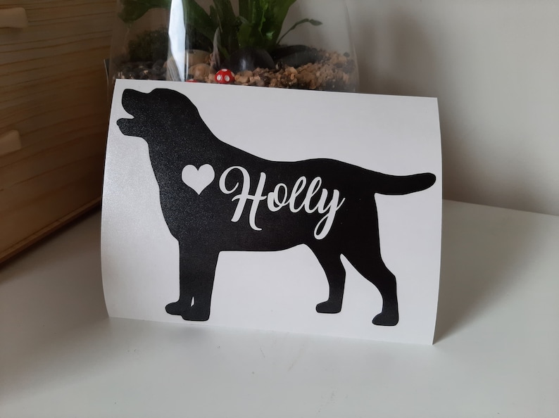 Op de afbeelding: Zwart Labrador Retriever silhouet met een wit hart en de naam "Holly" in een sierlijk lettertype. De sticker staat op een witte achtergrond. De hond kijkt naar links met zijn mond open.