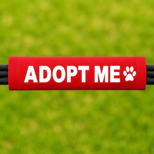 May include: A rectangular red fabric sleeve featuring the white text ADOPT ME and a white paw print graphic. The sleeve is positioned horizontally over two black cords, creating a clear and bold message for pet awareness and identification.