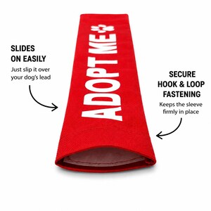 May include: A bright red rectangular dog leash sleeve featuring the white text ADOPT ME and a paw print icon. The sleeve includes text indicating it slides on easily over a dog's lead and utilizes a secure hook and loop fastening system.