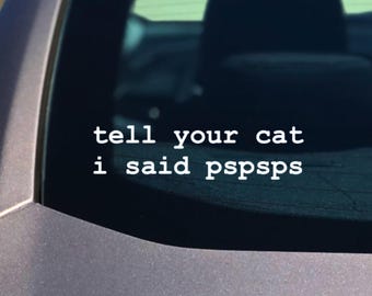 May include: A white vinyl decal applied to a dark car window featuring the text "tell your cat i said pspsps" in a simple, lowercase, sans-serif font. The sticker is positioned horizontally across the centre of the rear vehicle glass.