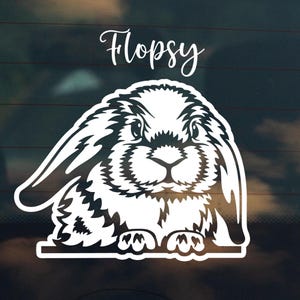 Puede incluir: Una calcomanía de vinilo blanco con una ilustración detallada de un conejo con orejas caídas. La palabra "Flopsy" está escrita encima del conejo en una fuente cursiva. El pelaje del conejo está representado con líneas intrincadas.