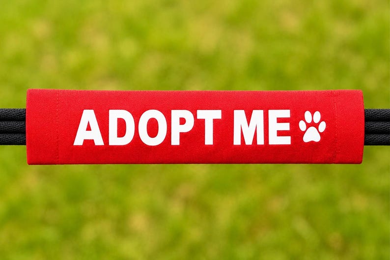 May include: A rectangular red fabric sleeve featuring the white text ADOPT ME and a white paw print icon. The sleeve is positioned horizontally and is threaded onto a black cord that extends from both the left and right sides of the fabric.