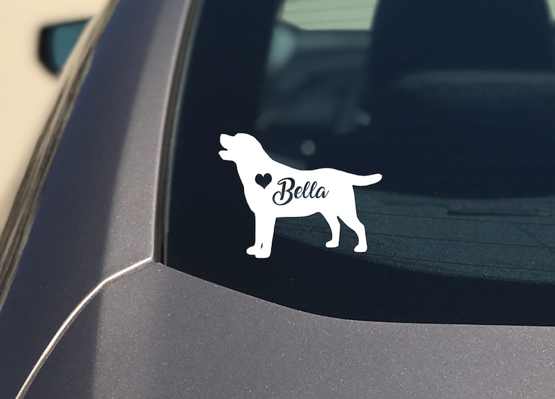 May include: White vinyl dog silhouette sticker with the name "Bella" and a heart. The sticker is displayed on a car window. A pet-themed car accessory.