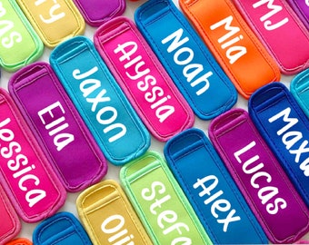 May include: A collection of colourful, personalised popsicle holders with names such as Jaxon, Ella, Noah, and Mia. The holders are made from a soft, neoprene-like material and are ideal for keeping popsicles cold and mess-free.
