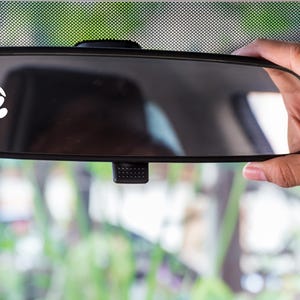 以下が含まれることがあります： 白いハートと足跡のステッカーが付いた車のバックミラー。 ミラーは手で持たれており、フロントガラスを通してぼやけた緑色の背景が見えます。 ステッカーはペットへの愛の象徴です。