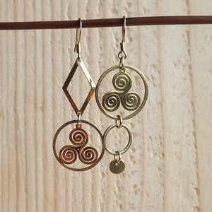 Op de afbeelding: Een paar gouden oorbellen met een diamantvorm en een cirkel met een drievoudig spiraalontwerp. De oorbellen hebben een kleine cirkel en een schijfcharme die eraan hangen. De oorbellen zijn op een houten achtergrond met de tekst "Clem et Tom".