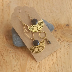 Op de afbeelding: Een paar gouden oorbellen met zwarte geometrische vormen en een ronde hanger. De oorbellen worden gepresenteerd op een bruine kaart met de tekst "Clem et Tom".