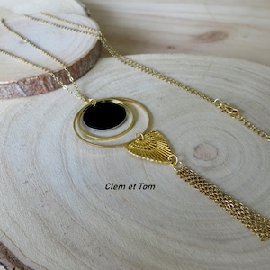 Peut inclure: Un collier en or avec un pendentif rond noir et un pompon doré. Le pendentif a un cercle plus petit à l'intérieur. Le collier est sur une surface en bois. Clem et Tom est écrit sur la surface.