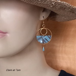 Peut inclure: Boucles d'oreilles créoles dorées avec un pendentif demi-cercle marbré bleu et blanc et une perle bleue en forme de larme suspendue en dessous.