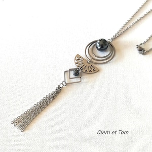 Peut inclure: Un collier pendentif argenté avec des éléments géométriques et en forme d'éventail. Le collier comprend une chaîne, deux cercles concentriques, un motif en forme d'éventail, un carré et une cascade de petites chaînes. Une perle gris foncé est incorporée. Le texte "Clem et Tom" est visible.