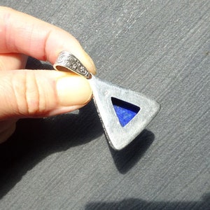 May include: Silver-toned pendant with a triangular shape and a vibrant blue stone inlay. The pendant has an ornate, detailed bail. The stone is a deep, rich blue color. The pendant is held in a hand.