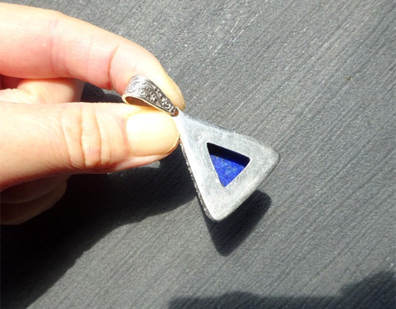 May include: Silver-toned pendant with a triangular shape and a vibrant blue stone inlay. The pendant has an ornate, detailed bail. The stone is a deep, rich blue color. The pendant is held in a hand.