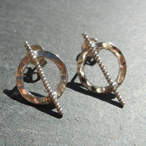 May include: A pair of silver earrings with a circle design and a textured bar that goes through the circle. The bar has a beaded texture.