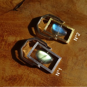 Op de afbeelding: Twee zilver- en goudkleurige hangers met rechthoekige frames, elk met een gepolijste, iriserende edelsteen. De edelstenen hebben blauwe en groene tinten. De hangers zijn gelabeld met "N°1" en "N°2" in witte tekst.