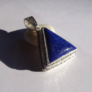 May include: A silver pendant with a triangular, deep blue lapis lazuli stone. The stone is set in a detailed silver frame with an ornate bail. The pendant is resting on a small, off-white stone, casting a shadow on a white surface.