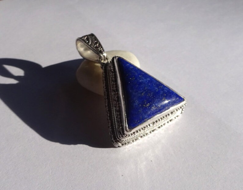 May include: A silver pendant with a triangular, deep blue lapis lazuli stone. The stone is set in a detailed silver frame with an ornate bail. The pendant is resting on a small, off-white stone, casting a shadow on a white surface.