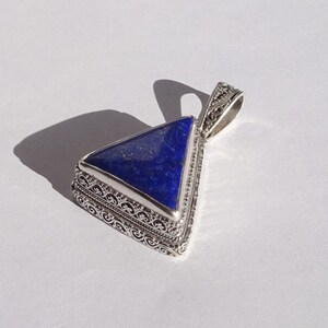 May include: A triangular pendant with a deep blue stone set in an ornate silver-colored metal frame. The pendant has a decorative bail for a chain. The design features detailed filigree work around the stone.